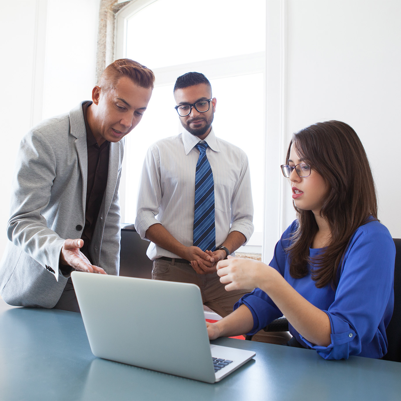 Three colleagues in a range of ages, genders, and races are seated closely behind a laptop in a bright office. One woman is using her pencil to point out something on the screen to her coworkers. 