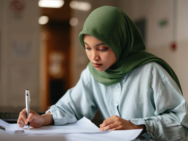A young woman in a hijba is writing on a piece of paper with a pen.