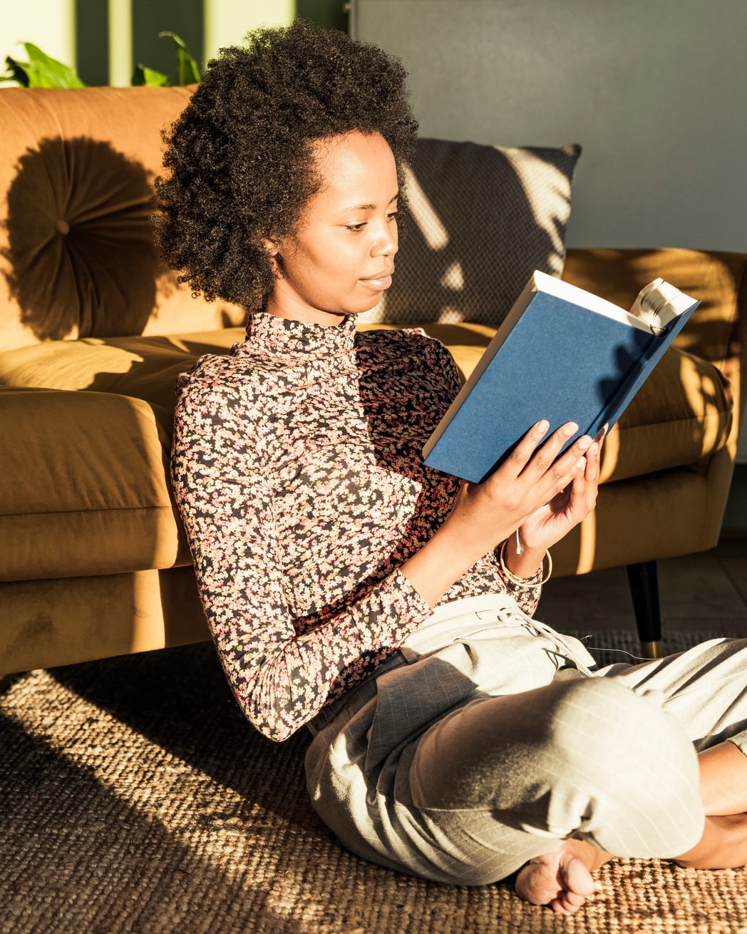 A woman is sitting on the floor, leaning against a leather couch. She is reading a book in a sunny room. 
