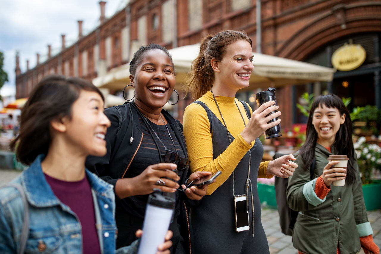 Happy multi-ethnic female friends with drinks walking on city street during vacation